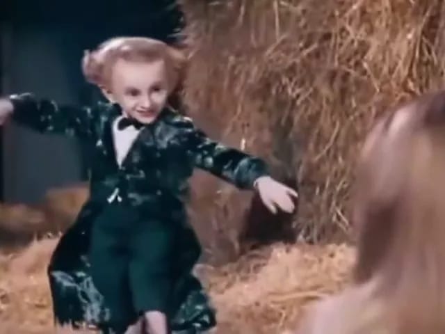 穿深色燕尾服的表演者在谷仓干草间张开双臂踮脚前进，前景里一名金发女子在看